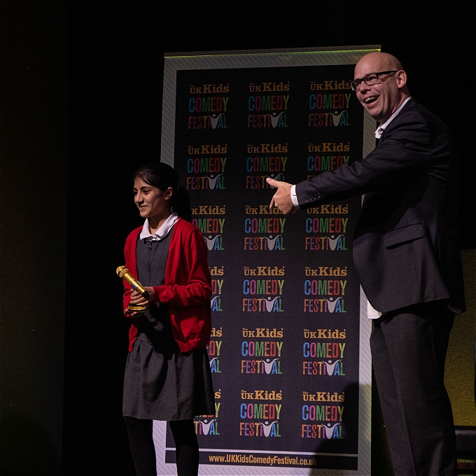 School pupil holding golden trophy on stage alongside comedian Rob Gee who is pointing at the winner