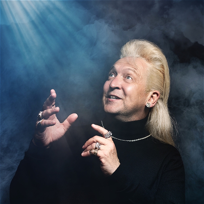 Male comedian with blonde hair, looking off into the distance, wearing a black jumper, with a blue background.