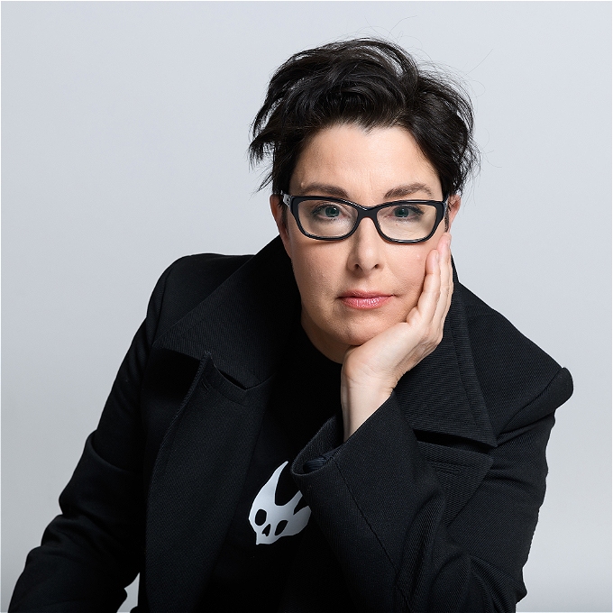 Female comedian with dark hair and glasses looking directly to camera, against a grey background.