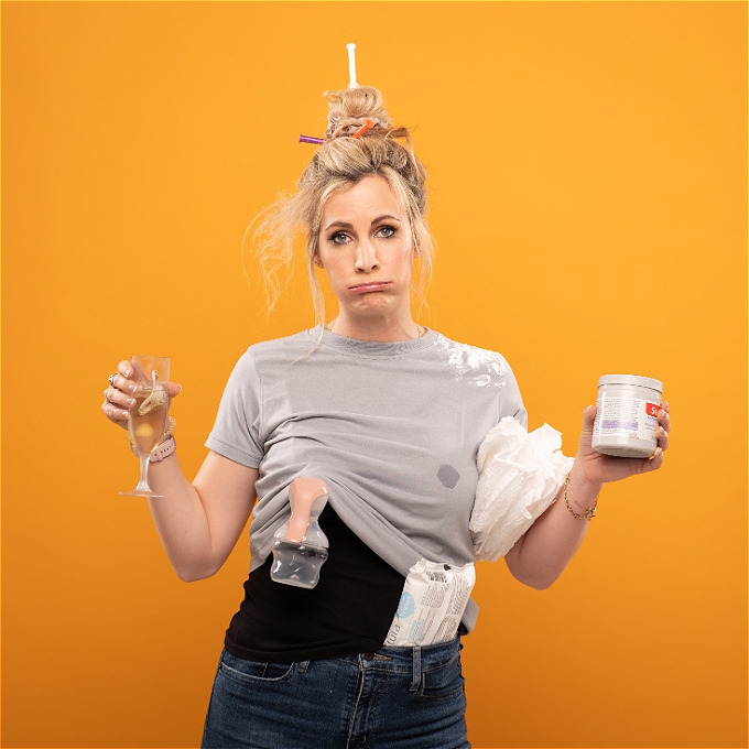 Hannah stands in front of a deep yellow background with an exhausted expression and her hair in disarray. She is holding nappy cream, prosecco with a dummy floating in it, nappies and has baby wipe tucked into her jeans.