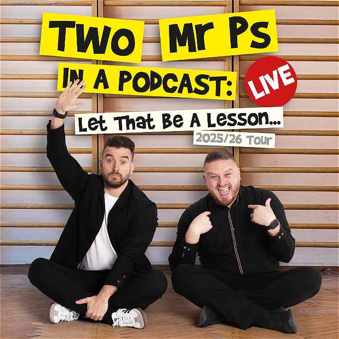 Two comedians sat on the floor in a school hall, one with their hand in the air