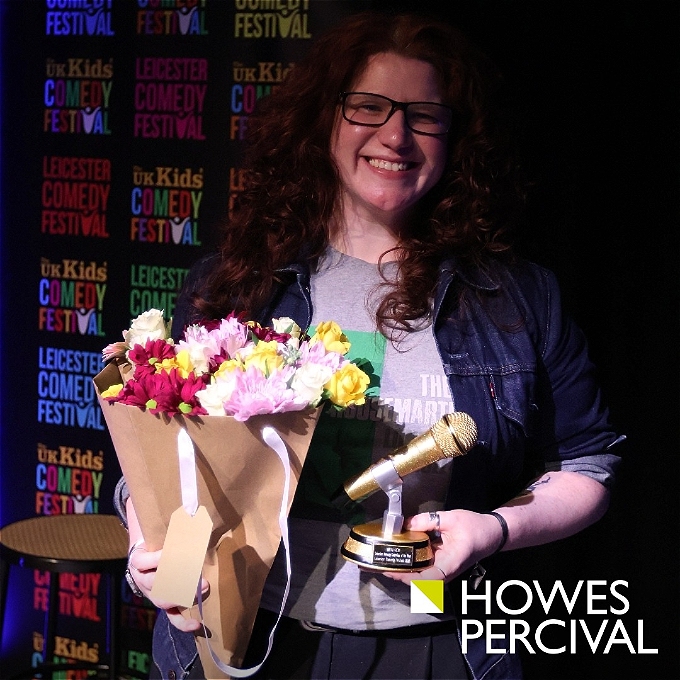 A female comedian holding a bunch of flowers and a trophy in the shape of a microphone. She has a big smile.