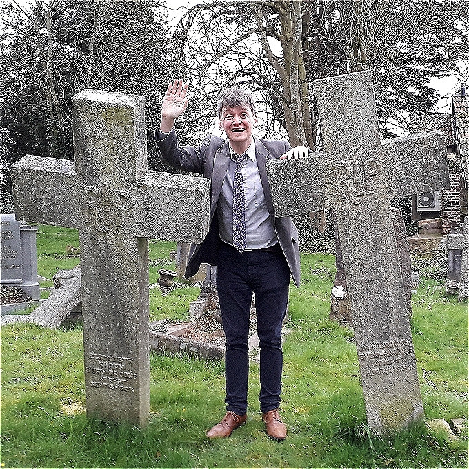 A young man dressed as an old man with grey hair in a battered suit and tie stands between two gravestones, waving cheerfully at the camera.
