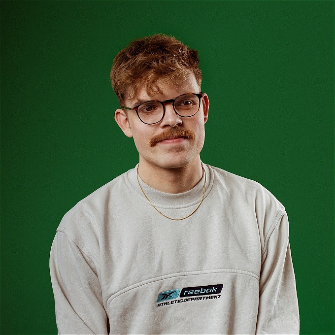 Male comedian with glasses sat down in front of a green backdrop