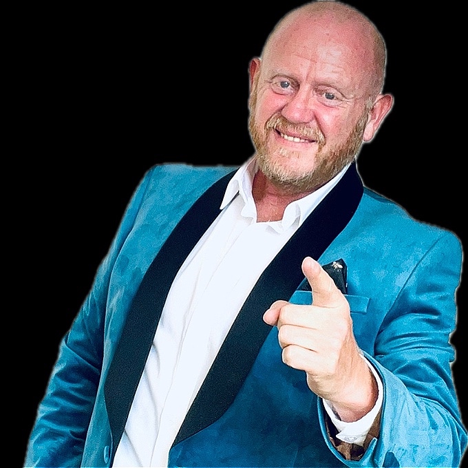 A Cheerful man with Bald head and reddish beard, dressed in a teal velvet jacket with black laps, points confidently toward the camera with a big smile.