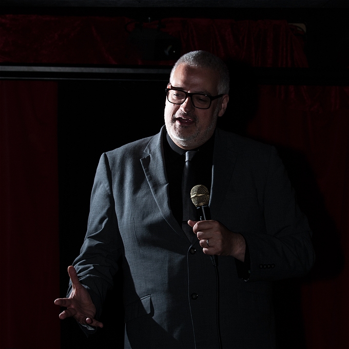 Male comedian in a suit with a mic in front of a dark background