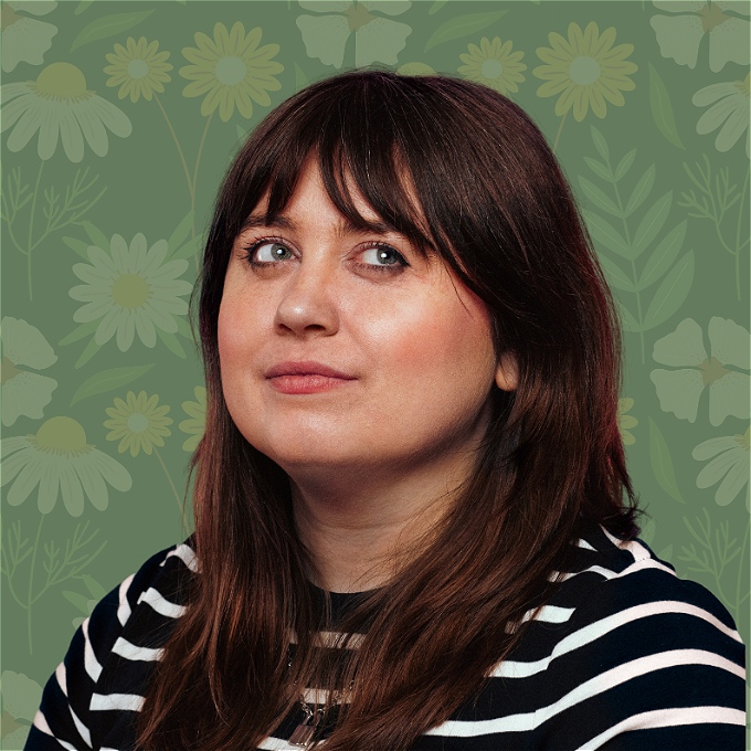 A white person with mid length reddish brown hair smiles whilst looking up on a green background with illustrated flowers