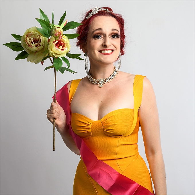 Female comedian with red hair smiling in a yellow dress, holding a bunch of flowers