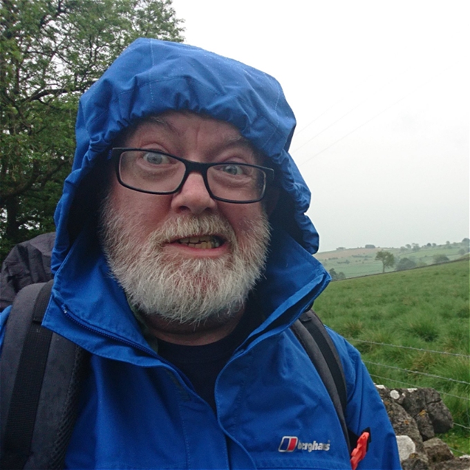 Man with beard and glasses wearing blue cagoule standing by a field
