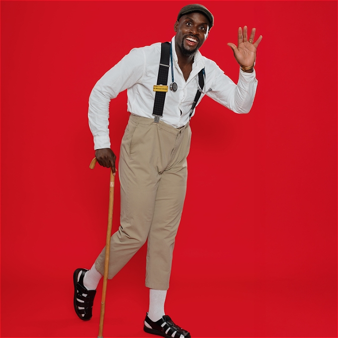 Gentleman smiling sporting a flat cap, braces with a stethoscope around his neck and a walking stick in his right hand.