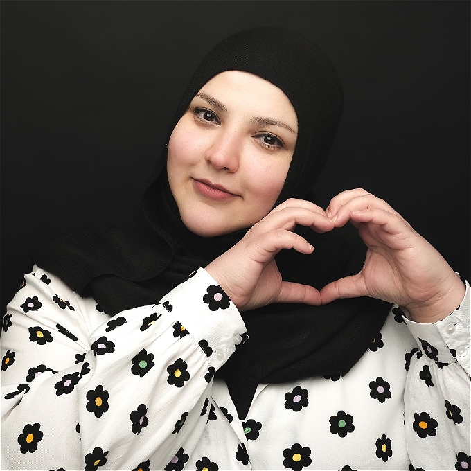Female comedian making a heart shape with her hands, and wearing a flowery shirt, on a black background.