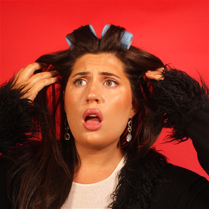 White female comedian with brown hair in big curlers. She wears a white shirt with a black fur trimmed cardigan