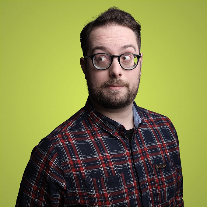A male comedian with brown hair and thick glasses, a short beard and a red checked shirt stands in front of a green background. He is stood at a 45 degree angle towards the right but his eyes are looking the opposite direction.