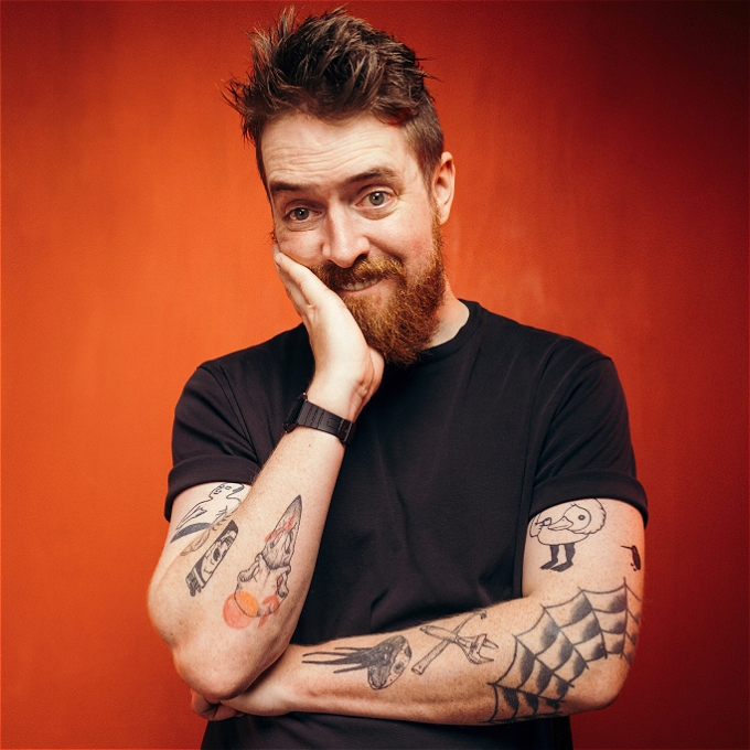 Male comedian has head in hands looking perplexed against an orange background