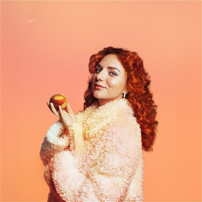 Female comedian wearing a fluffy white jackets smirks while holding a peach