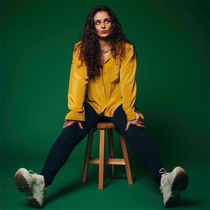 Comedian Farah Sharp sat on a stool in front of a green background looking perplexed