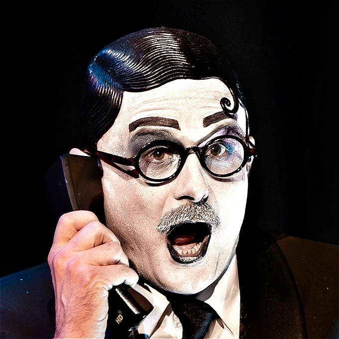 Male comedy character with white face make up, black rubber wig and round glasses, holds a telephone to their ear and looks surprised.