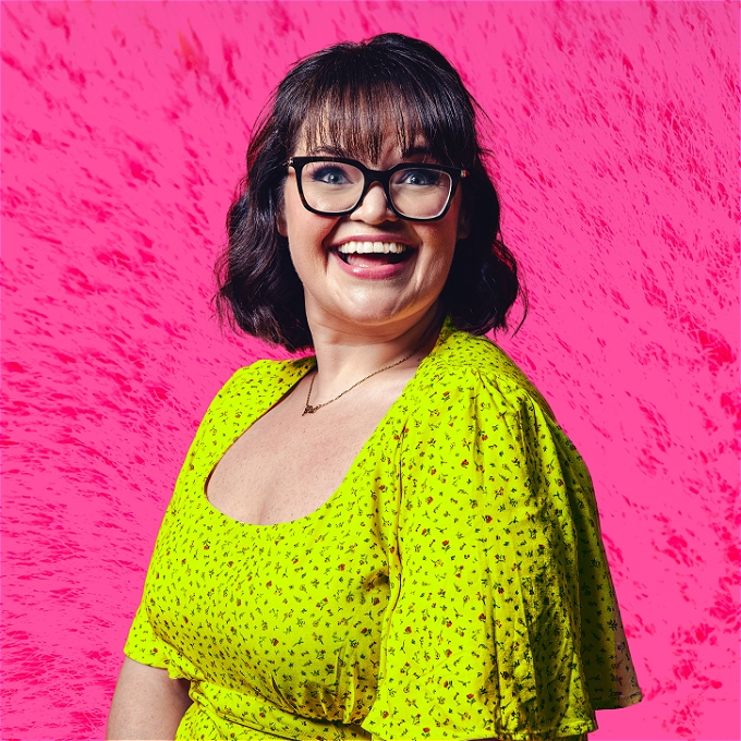 Female comedian with black hair and glasses against a bright pink background