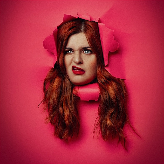 Female comedian with ginger hair pushing through a pink piece of paper, making a funny face to camera
