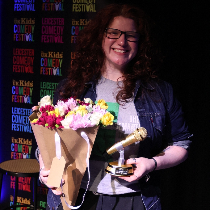 A female comedian holding a bunch of flowers and a trophy in the shape of a microphone. She has a big smile.