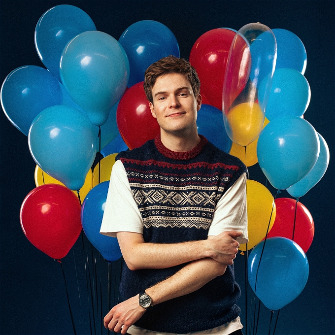 A male comedian smiles wryly in front lots of balloons
