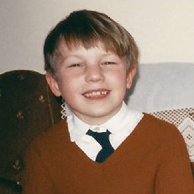 Young boy in the 1970s with short brown hair dressed in a red v-necked jumper and a black tie