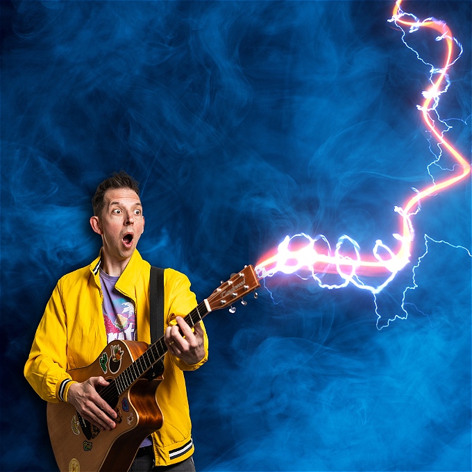 Male comedian is stood looking shocked holding a guitar with a lightening bolt coming out of it