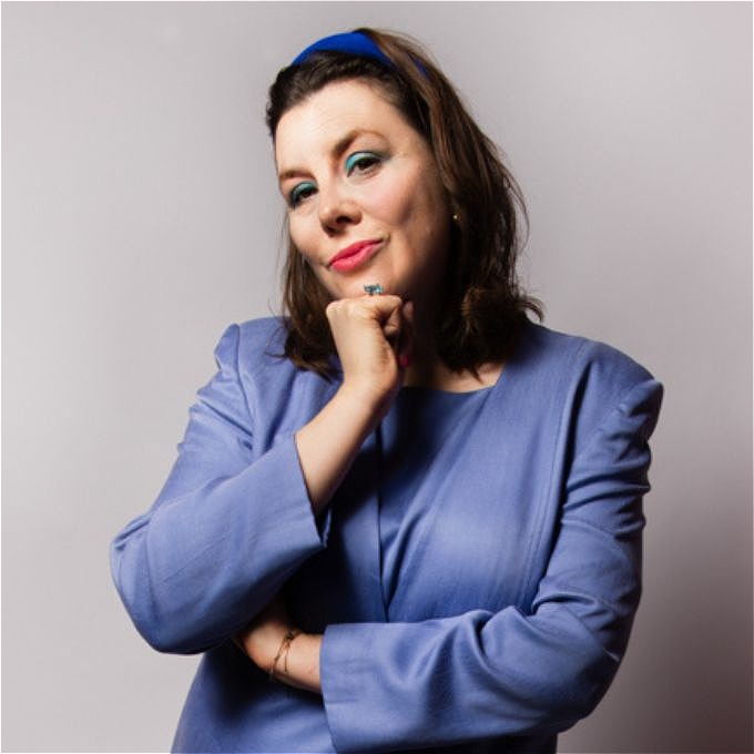 Caucasion female comedian with brown hair wearing a blue headband and lilac suit from the 90s