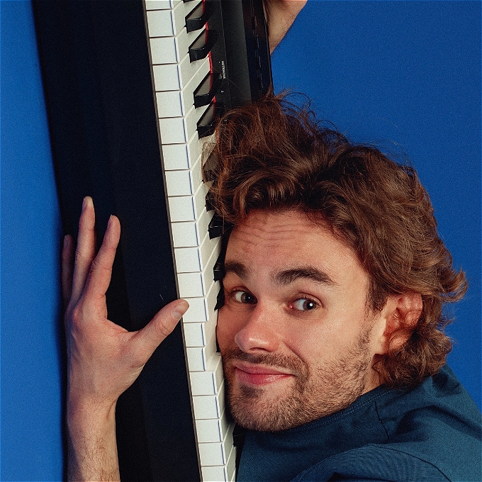Male comedian with mid-length brown hair and a light beard with his head against a keyboard. He is clutching the keyboard with his hand and looking directly into the camera.