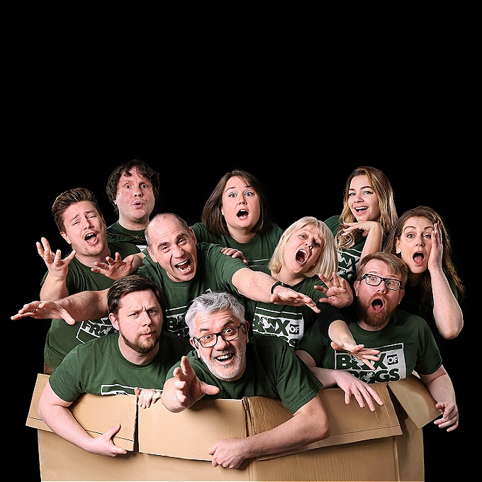Group of 10 Improvisers dressed in green T shirts laughing and reaching out from inside a large cardboard box