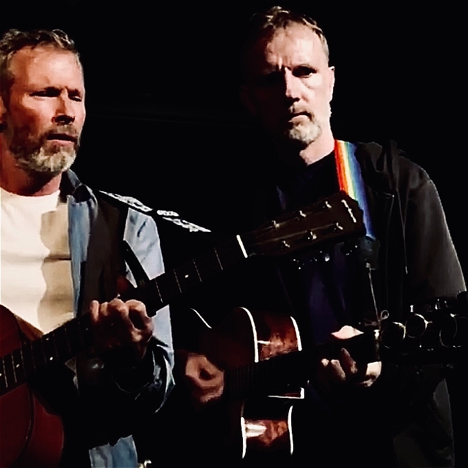Two Male Musical comedians standing side by side playing guitars