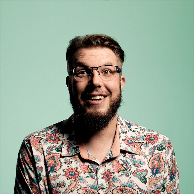 Male comedian with brown hair, brown beard and blue glasses wearing a paisley shirt in front of a green background