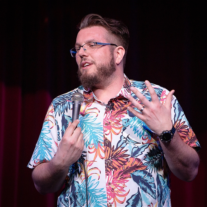 Male comedian with brown hair, brown beard and blue glasses wearing a paisley shirt in front of red background