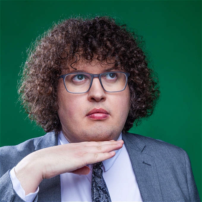 A curly haired bespectacled male comedian looks up and away.