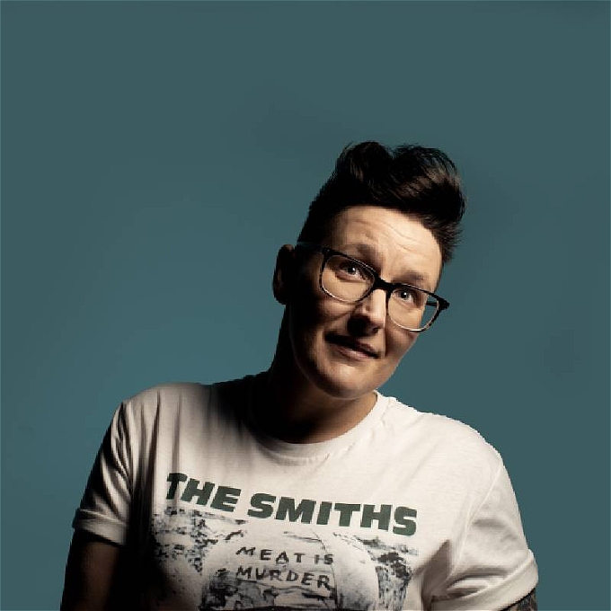 Female comedian with short brown hair and glasses, wearing a white Smiths t shirt, looking upwards, on a blue green background