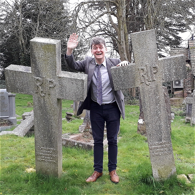 A young man dressed as an old man with grey hair in a battered suit and tie stands between two gravestones, waving cheerfully at the camera.