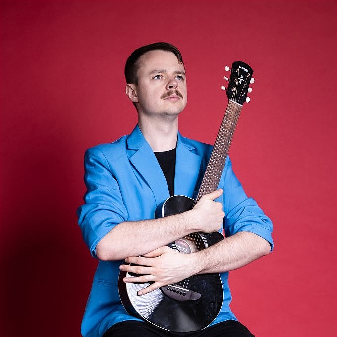 Male comedian with brown hair, blue jacket and a black guitar