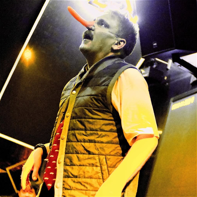 Man wearing a long fake nose looking towards the sky. He's wearing a gilet and a shirt and tie.