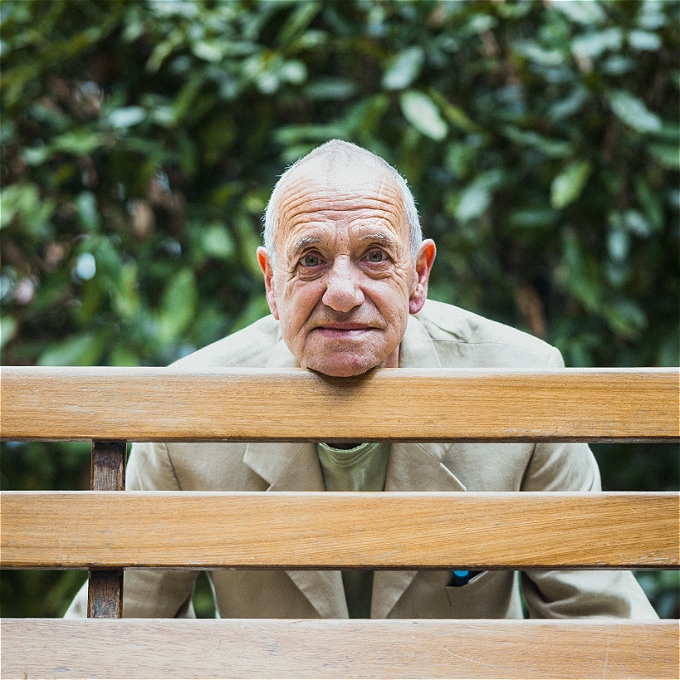 A male comedian in a beige suit jacket and green t-shirt rests his chin on the back of a wooden bench. A shrub is in the background.