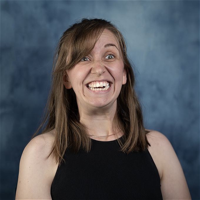 Female comedian smiling a big toothy smile with her eye wide in front of a blue background