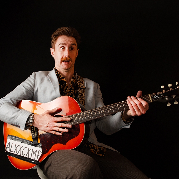 Male comedian with red hair holding a red sunburst guitar and leaning back on a chair with a shocked expression on his face