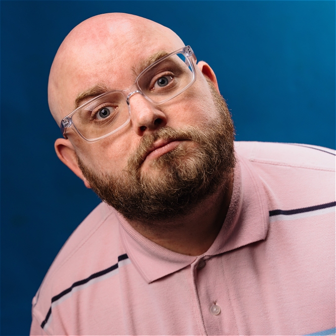Will Duggan a bald comdian with clear glasses is looking at the camera in a pink polo shirt