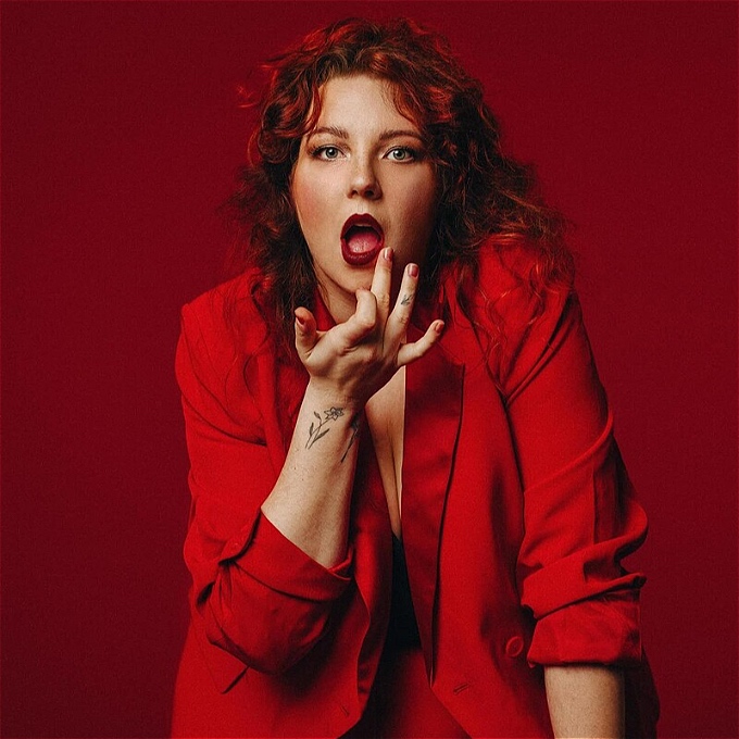 Female comedian with red hair, wearing a red suit, removes something from the side of her mouth.