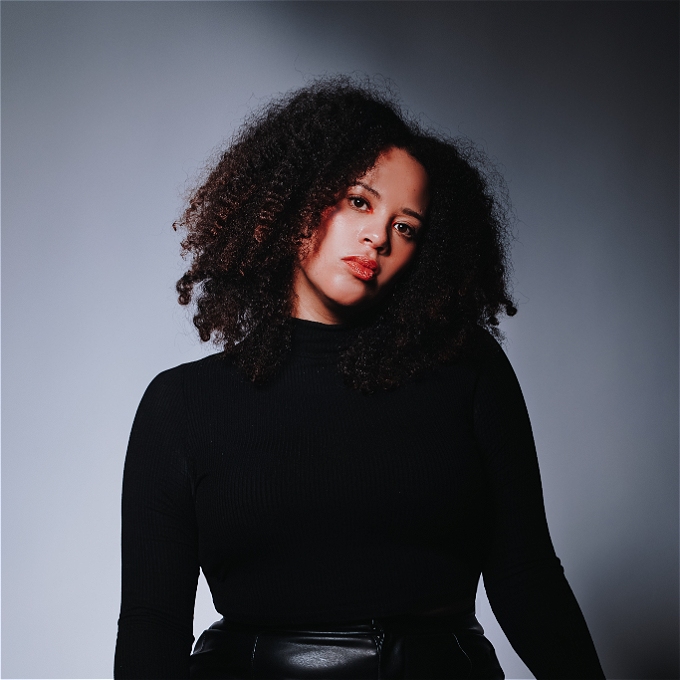 Mixed race female comedian wearing black turtleneck, with curly hair