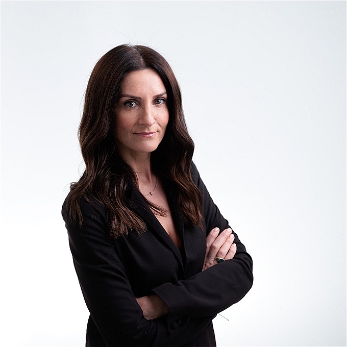 female comedian with brown hair wearing a black jacket with arms folded