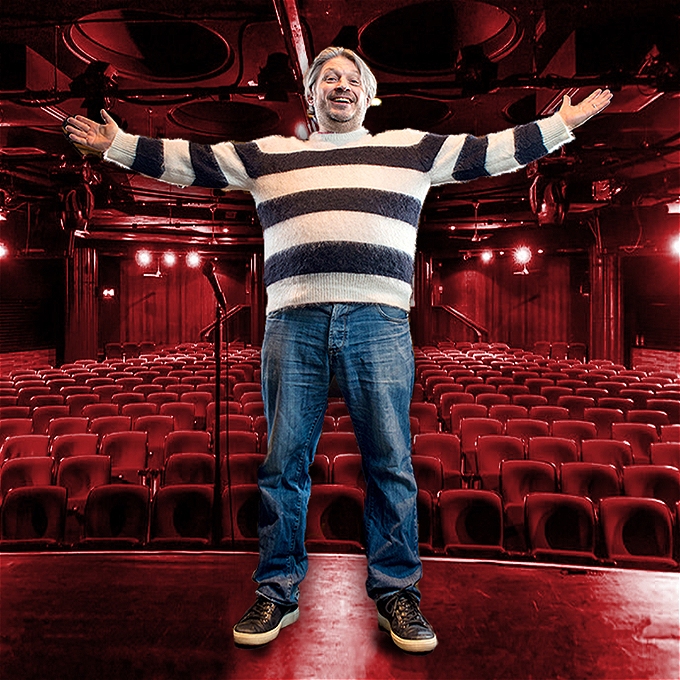Richard Herring stands with his arms outstretch with his back to a theatre auditorium