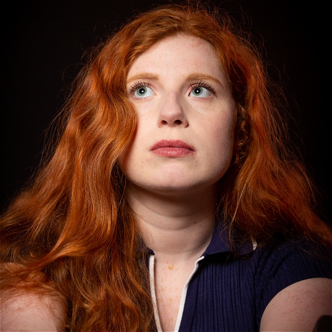 Female comedian with red hair looks up on dark background