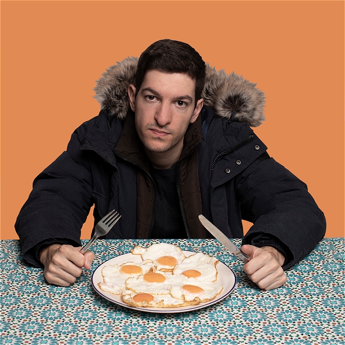 Male Comedian sitting at a table with an art deco table cloth and a plate of six eggs