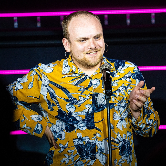 Comedian Andrew White stands at a microphone smiling in a floral yellow shirt and gold hoop earrings