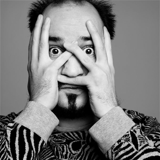 White male comedian with short brown hair and a zebra patterned jumper holding his hands to his face, his eyes can be seen inbetween his fingers
